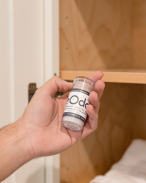 Hand reaching for Tobacco Vanilla DeOdor from a bathroom cabinet — warm light on wood shelves and folded white towels
