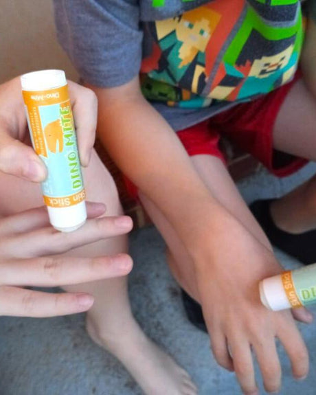 Kids applying open Dino-Mite Skin Stick solid lotion to knuckles outdoors — in-use sensory moment