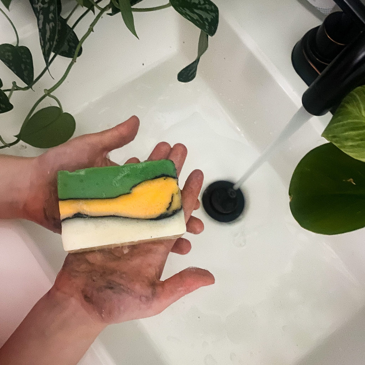 Soiled hands hold layered green-and-yellow Dino-Mite soap over a white sink with running water