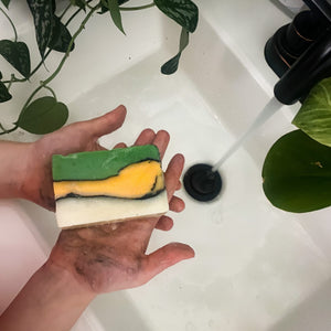 Soiled hands hold layered green-and-yellow Dino-Mite soap over a white sink with running water