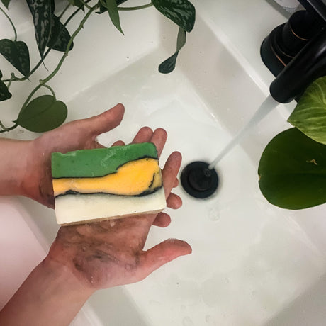Soiled hands hold layered green-and-yellow Dino-Mite soap over a white sink with running water
