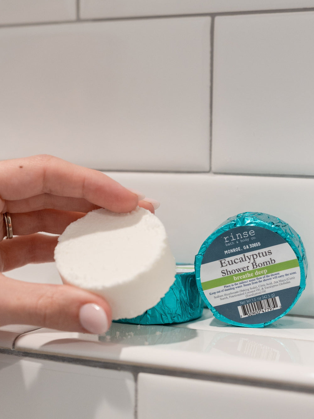 Hand presenting a powdery Eucalyptus shower bomb with teal wraps behind — white subway tile catching soft bathroom light