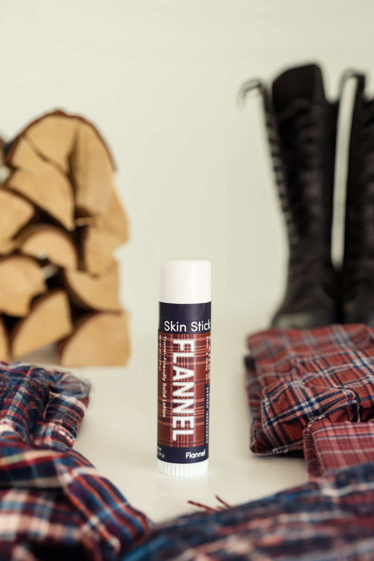 Flannel solid lotion stick standing against a plaid flannel shirt, leather boots and split firewood — cedar, clove and sweet tobacco, the men's gift that smells like a cabin porch. Rinse Bath & Body Co.