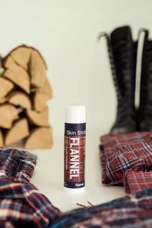 Flannel solid lotion stick standing against a plaid flannel shirt, leather boots and split firewood — cedar, clove and sweet tobacco, the men's gift that smells like a cabin porch. Rinse Bath & Body Co.