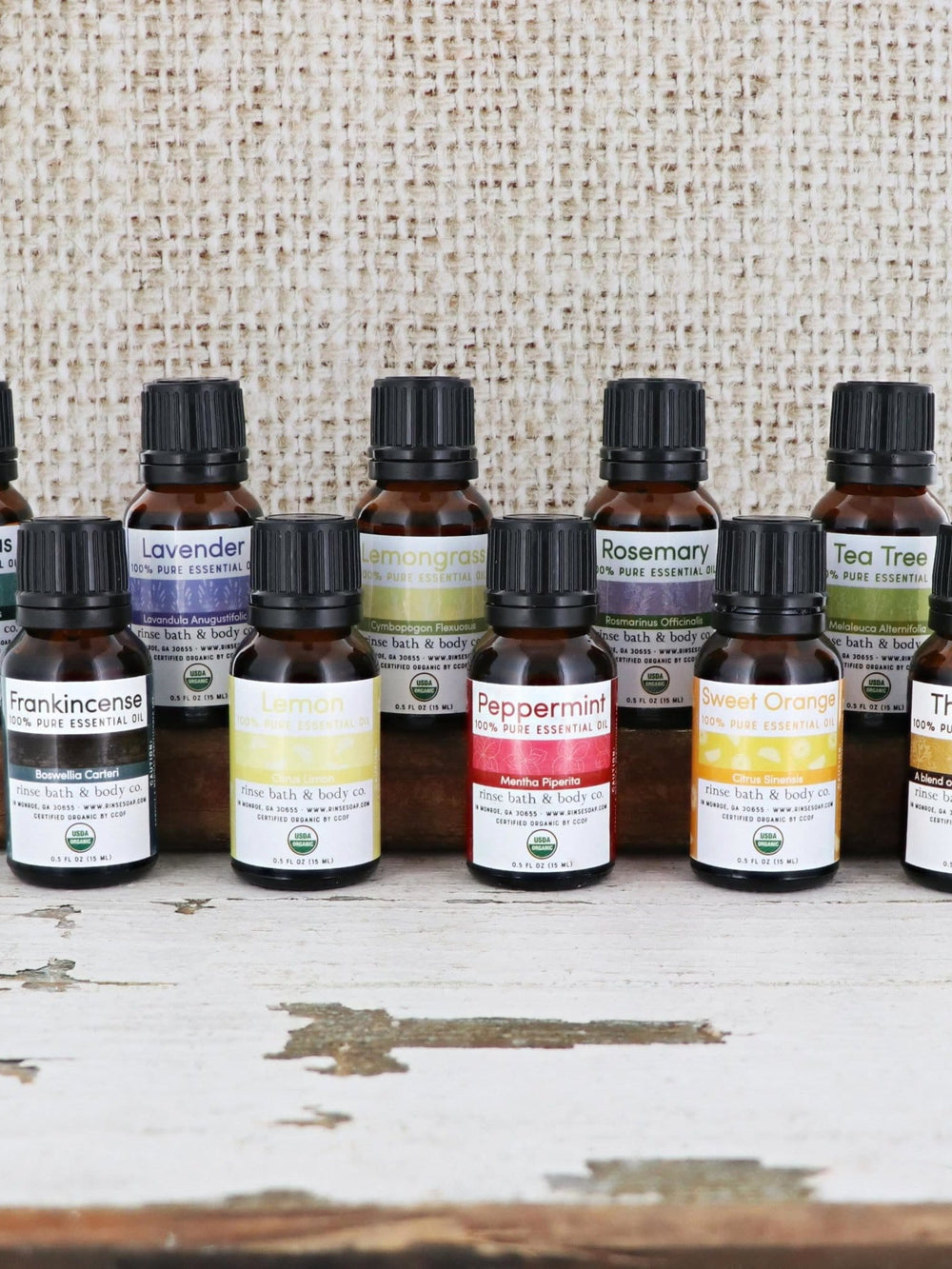 Ten amber essential oil bottles lined on a weathered wood beam — colorful USDA Organic labels against rustic burlap