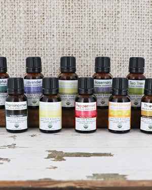 Ten amber essential oil bottles lined on a weathered wood beam — colorful USDA Organic labels against rustic burlap