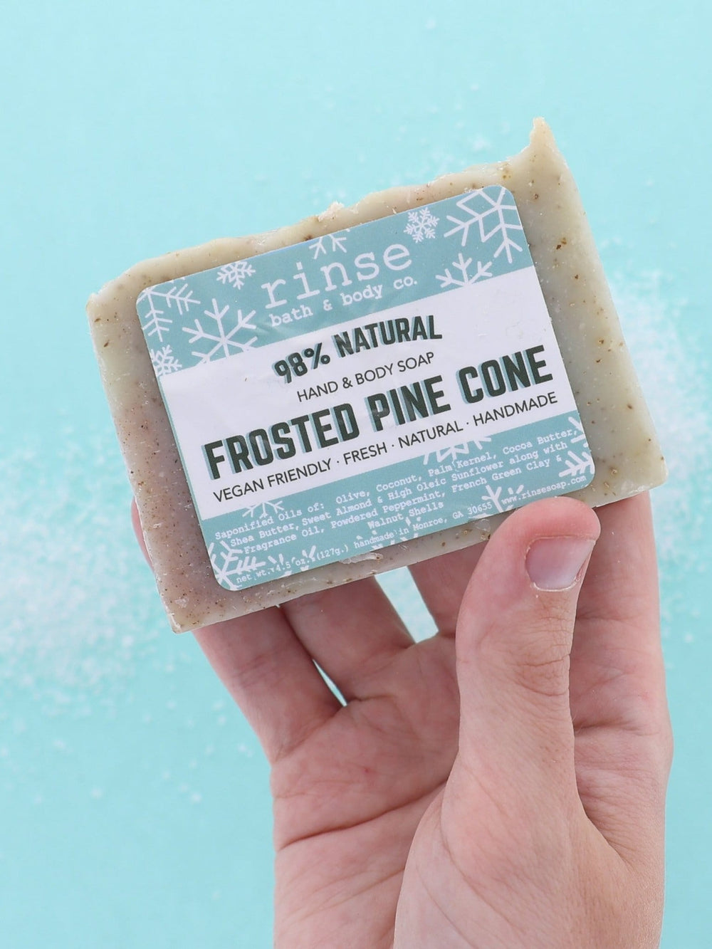 Hand holding Frosted Pine Cone soap over icy blue with white crystal grains — frosty sparkle in bright light