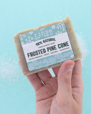 Hand holding Frosted Pine Cone soap over icy blue with white crystal grains — frosty sparkle in bright light