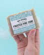 Hand holding Frosted Pine Cone soap over icy blue with white crystal grains — frosty sparkle in bright light