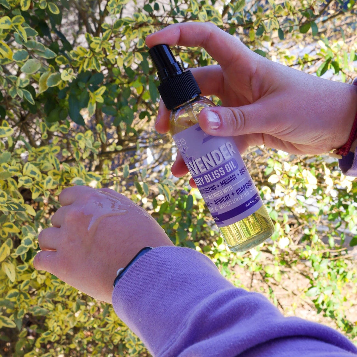 Hands misting Lavender Body Bliss Oil onto skin outdoors — purple sleeve and variegated foliage in sun