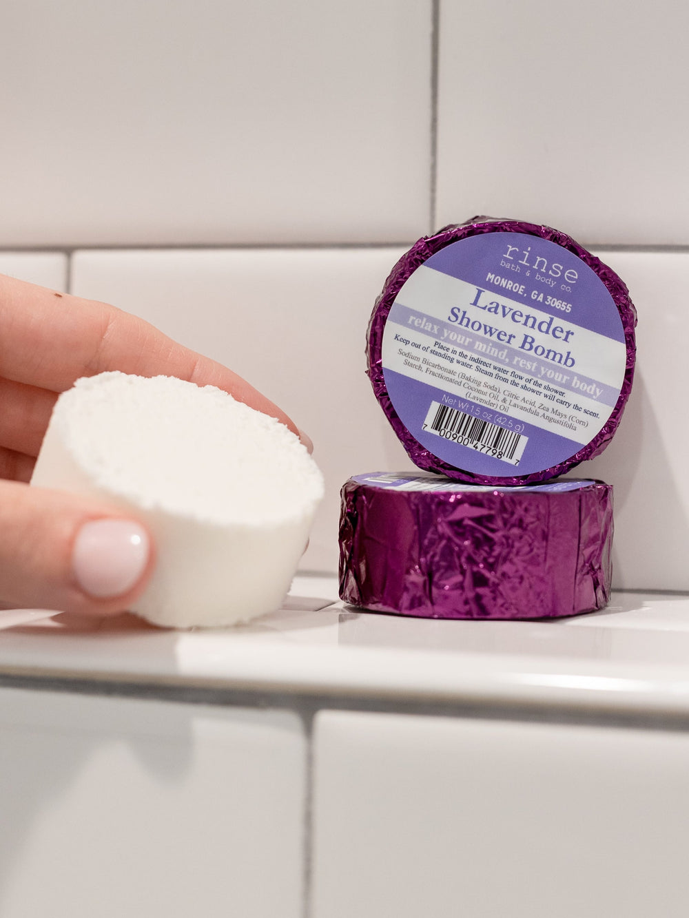Unwrapped Lavender Shower Bomb held before subway tile — purple foil pack resting on the white ledge