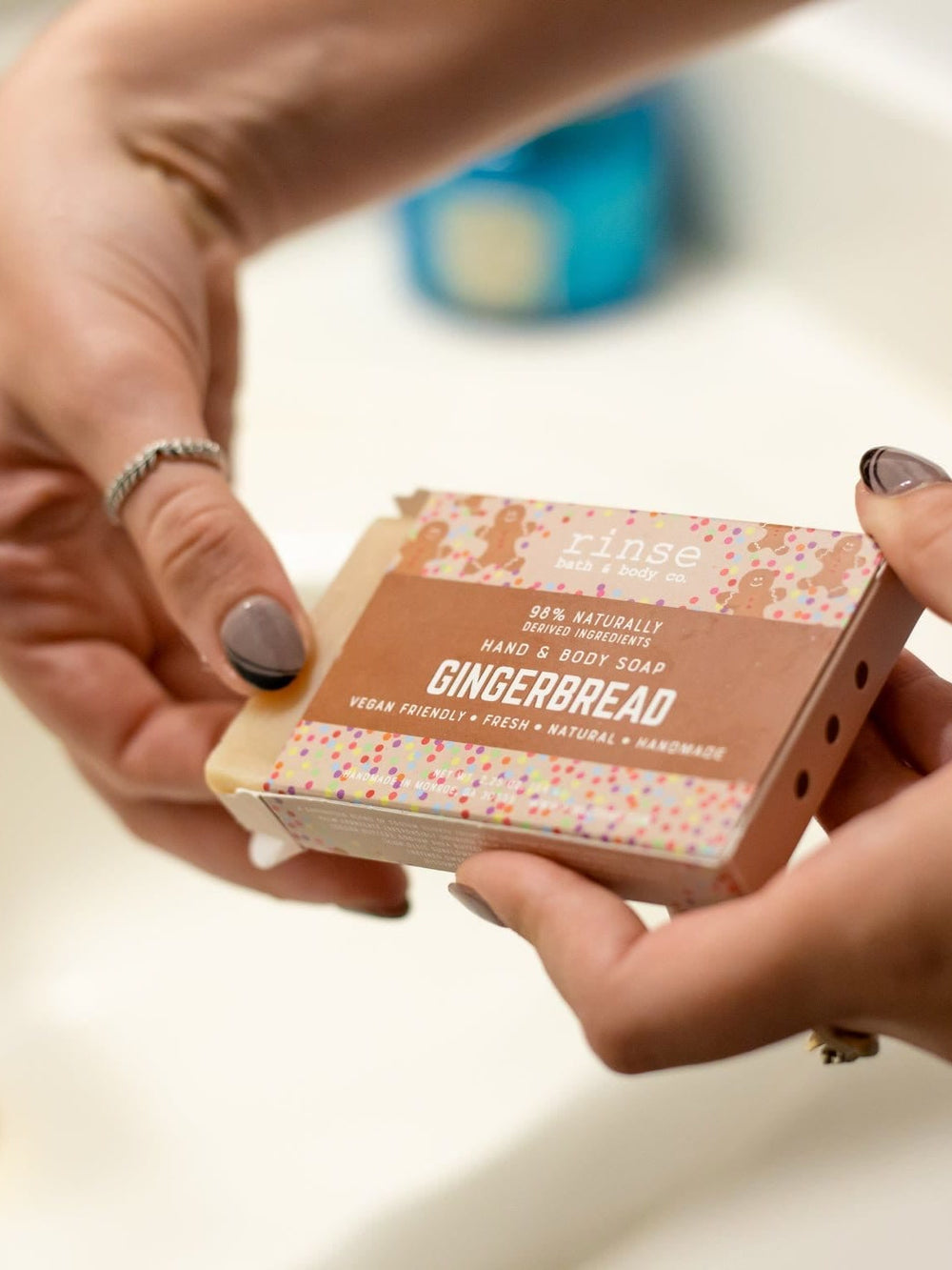 Hands sliding Gingerbread soap from its brown sleeve over a white sink — gold faucet soft in bathroom light