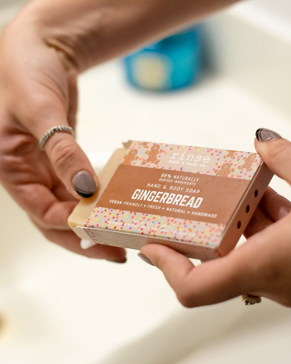 Hands sliding Gingerbread soap from its brown sleeve over a white sink — gold faucet soft in bathroom light