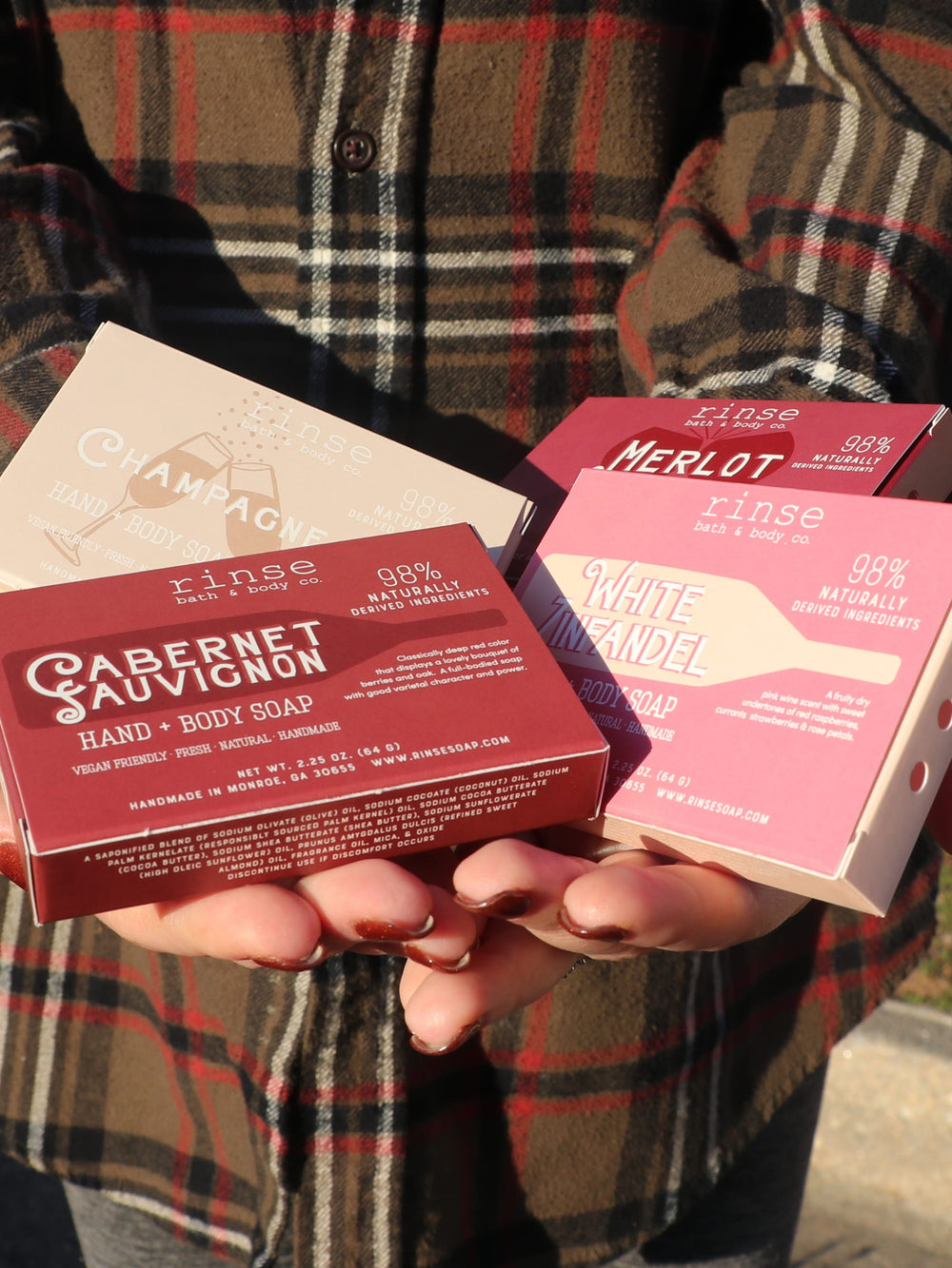 Hands cradling four wine-scented mini soap boxes — Cabernet Merlot Champagne and White Zin against red plaid in sun