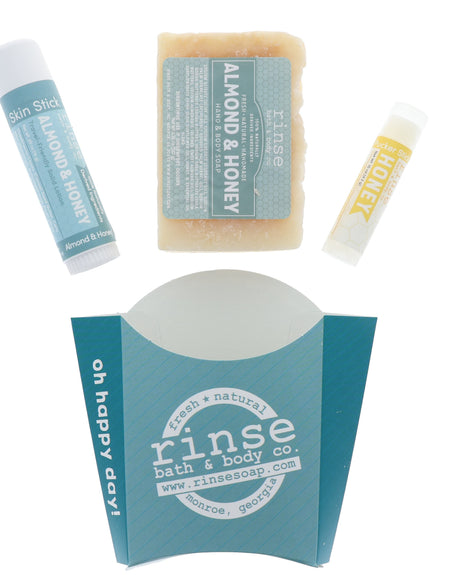 The Almond & Honey set from the everyday assortment laid out flat — bar soap, solid lotion skin stick and Honey lip balm above the teal carton. Rinse Bath & Body Co.