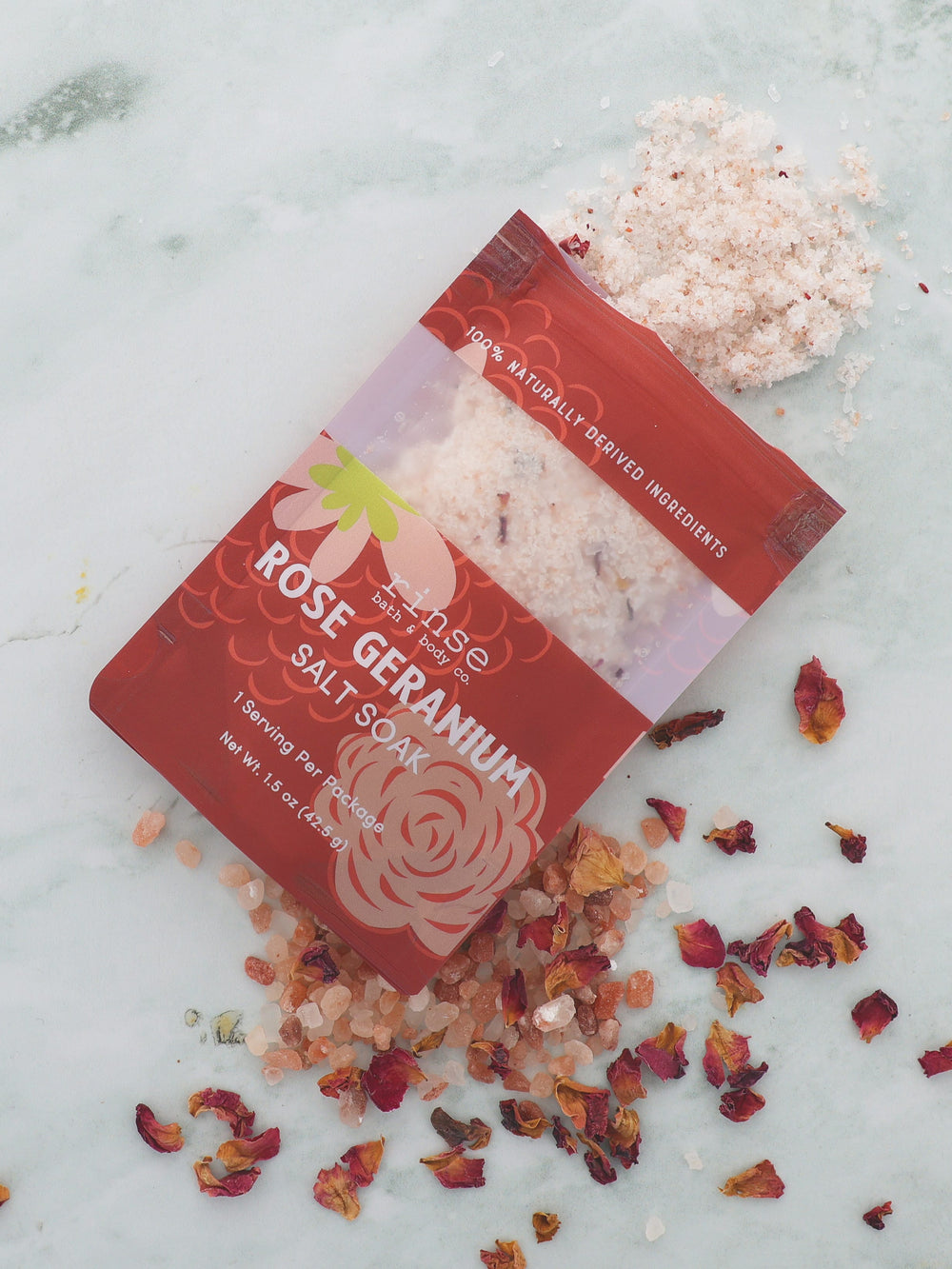 Rose Geranium Salt Soak pouch opened on veined marble — pink crystals and rose petals spilling in soft daylight