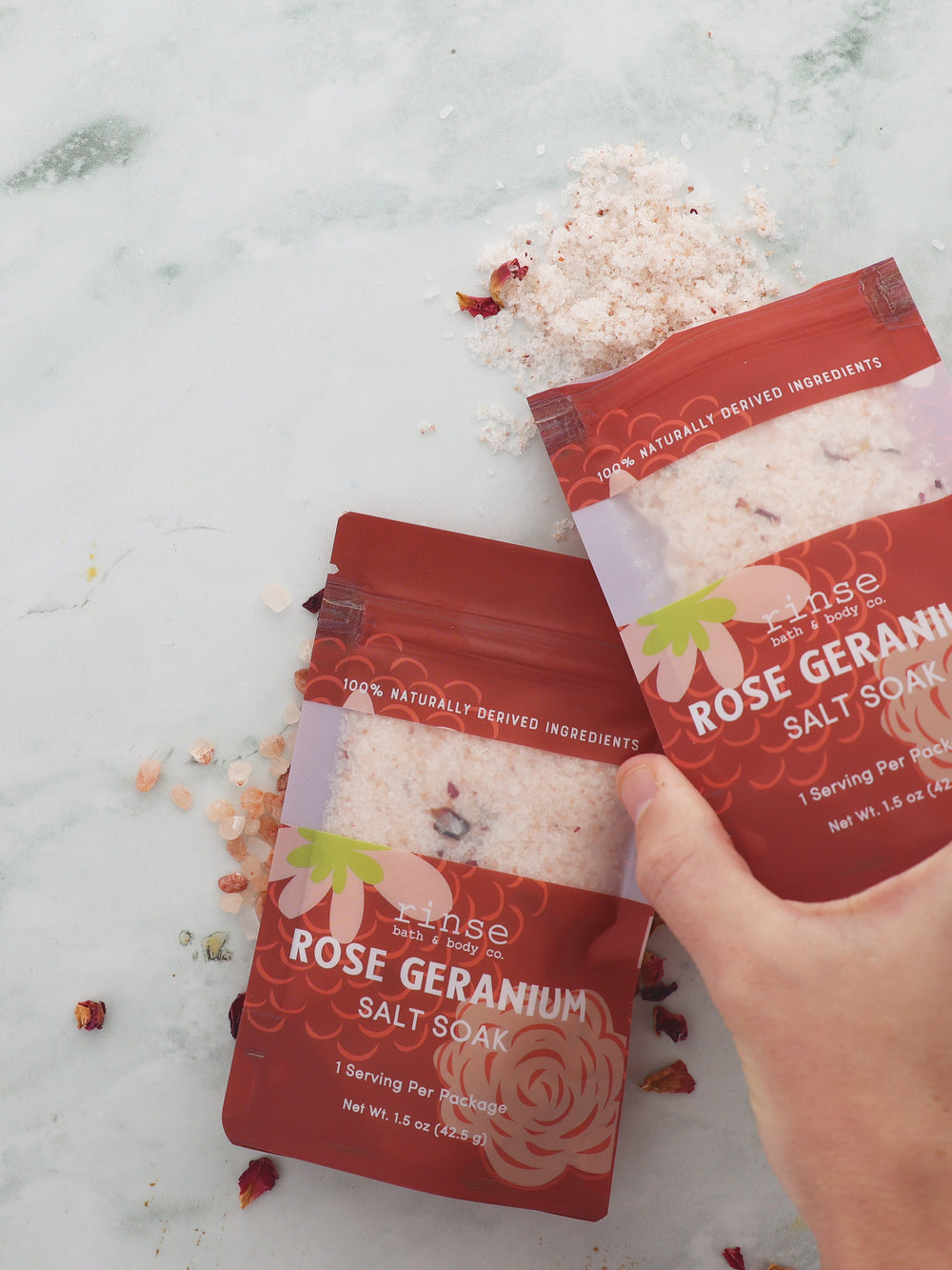Hand tilting Rose Geranium Salt Soak onto white marble — pink salts and dried petals cascading from the terracotta pouch