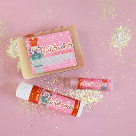 The Nutcracker gift set on pink paper scattered with gold glitter — Nutcracker bar soap, solid lotion skin stick and lip balm. Rinse Bath & Body Co.