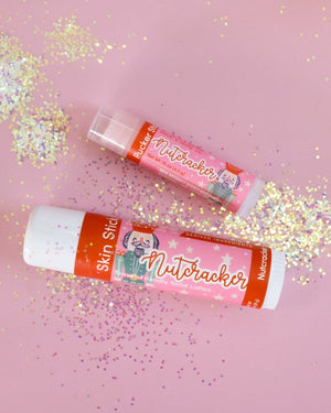 Nutcracker Skin Stick lifestyle on pastel pink scattered with iridescent glitter — solid lotion in-use mood