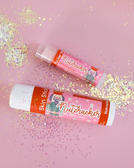 Nutcracker Skin Stick lifestyle on pastel pink scattered with iridescent glitter — solid lotion in-use mood