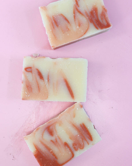 Three Peach soap bars scatter on matte light pink — cream and peach-orange swirls in soft flat-lay light