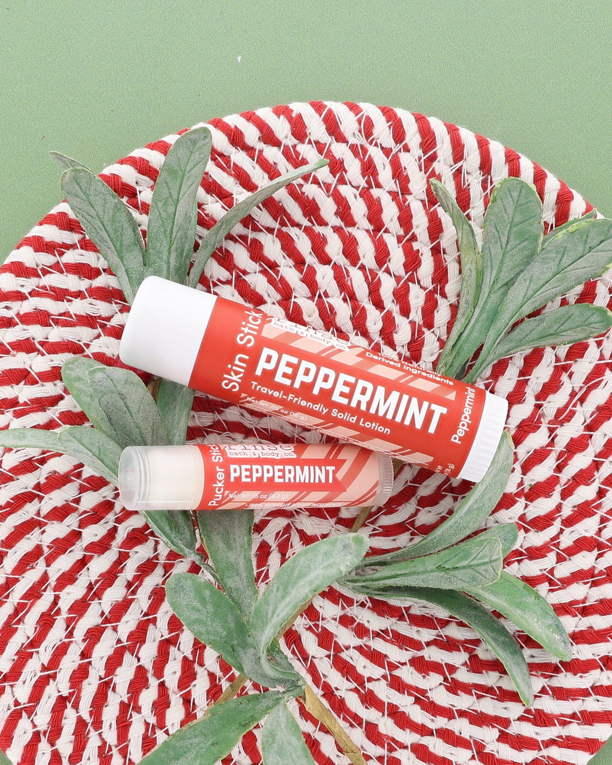 Peppermint Skin Stick and Peppermint Pucker Stick on a red and white woven plate with a frosted greenery sprig