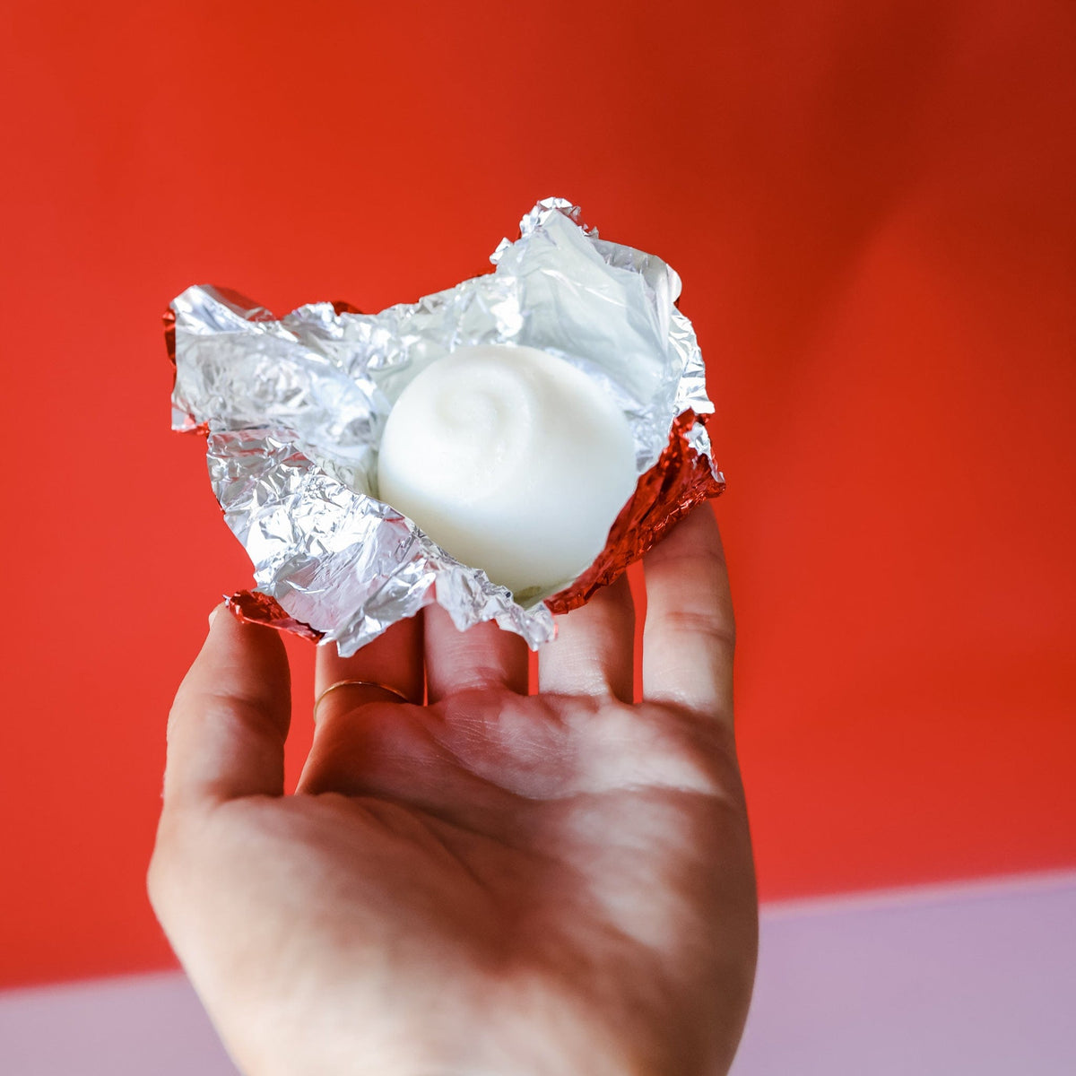 Hand presenting an unwrapped white Peppermint Tub Truffle in opened red foil — swirl top against bold red and lavender