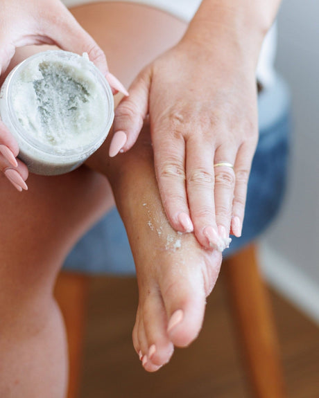 A woman scooping Peppofoot Polish and working it over her foot — gritty pumice and cooling peppermint, the pre-pedicure step done at home. Rinse Bath & Body Co.