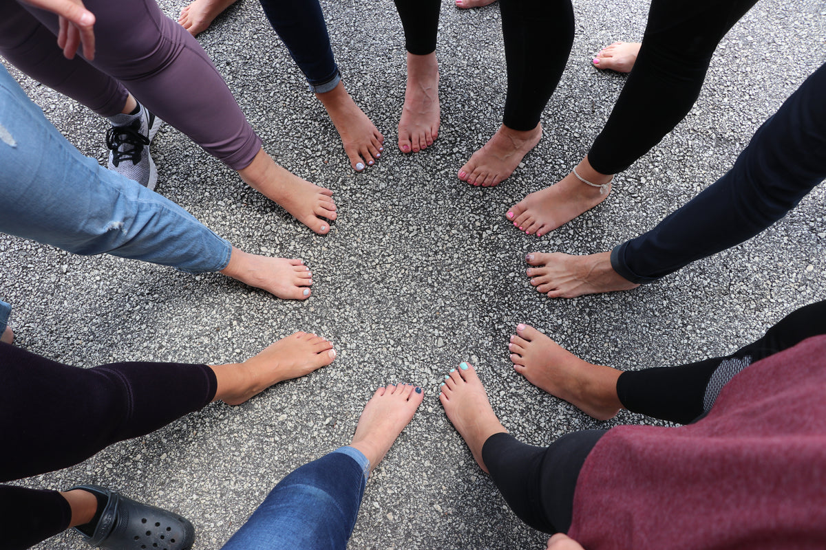 Five pairs of bare feet arranged in a circle on pavement, toes in — the after-the-hike moment Peppofoot spray is made for. Rinse Bath & Body Co.