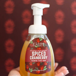 Hand holding a Spiced Cranberry Foaming Hand Soap pump bottle against a deep red patterned wall — cranberry label over amber castile soap