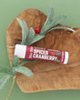 Spiced Cranberry Skin Stick on an olive wood board with a frosted greenery sprig and red berry on sage green