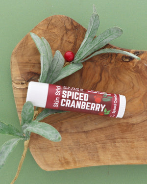 Spiced Cranberry Skin Stick on an olive wood board with a frosted greenery sprig and red berry on sage green
