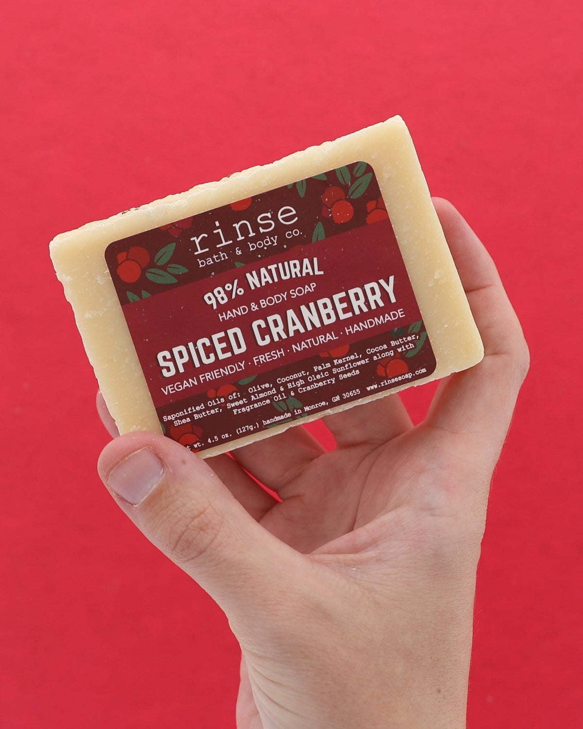 Hand holding Spiced Cranberry soap against solid red — seed-flecked cream bar in use in warm studio light