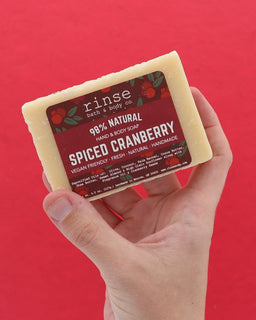 Hand holding Spiced Cranberry soap against solid red — seed-flecked cream bar in warm studio light