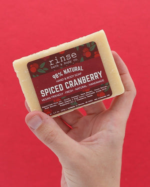 Hand holding Spiced Cranberry soap against solid red — seed-flecked cream bar in warm studio light