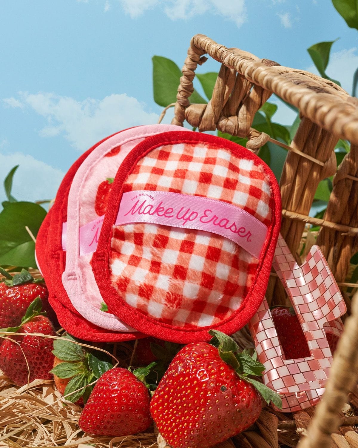 Stack of strawberry-print and gingham MakeUp Eraser cloths beside pink hair clip on straw with fresh strawberries outdoors