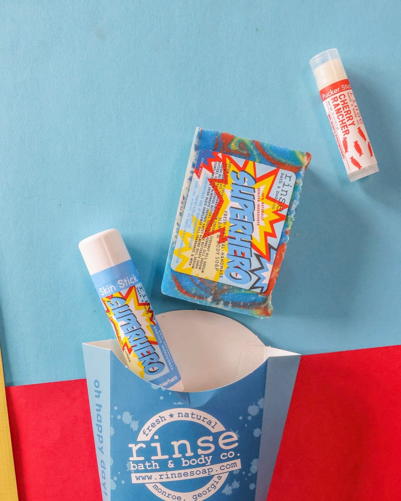 The Superhero kids gift set styled on blue, red and yellow colour-blocked paper — rainbow-swirl bar soap, Superhero solid lotion skin stick and a Cherry Rancher lip balm beside the blue carton. Rinse Bath & Body Co.
