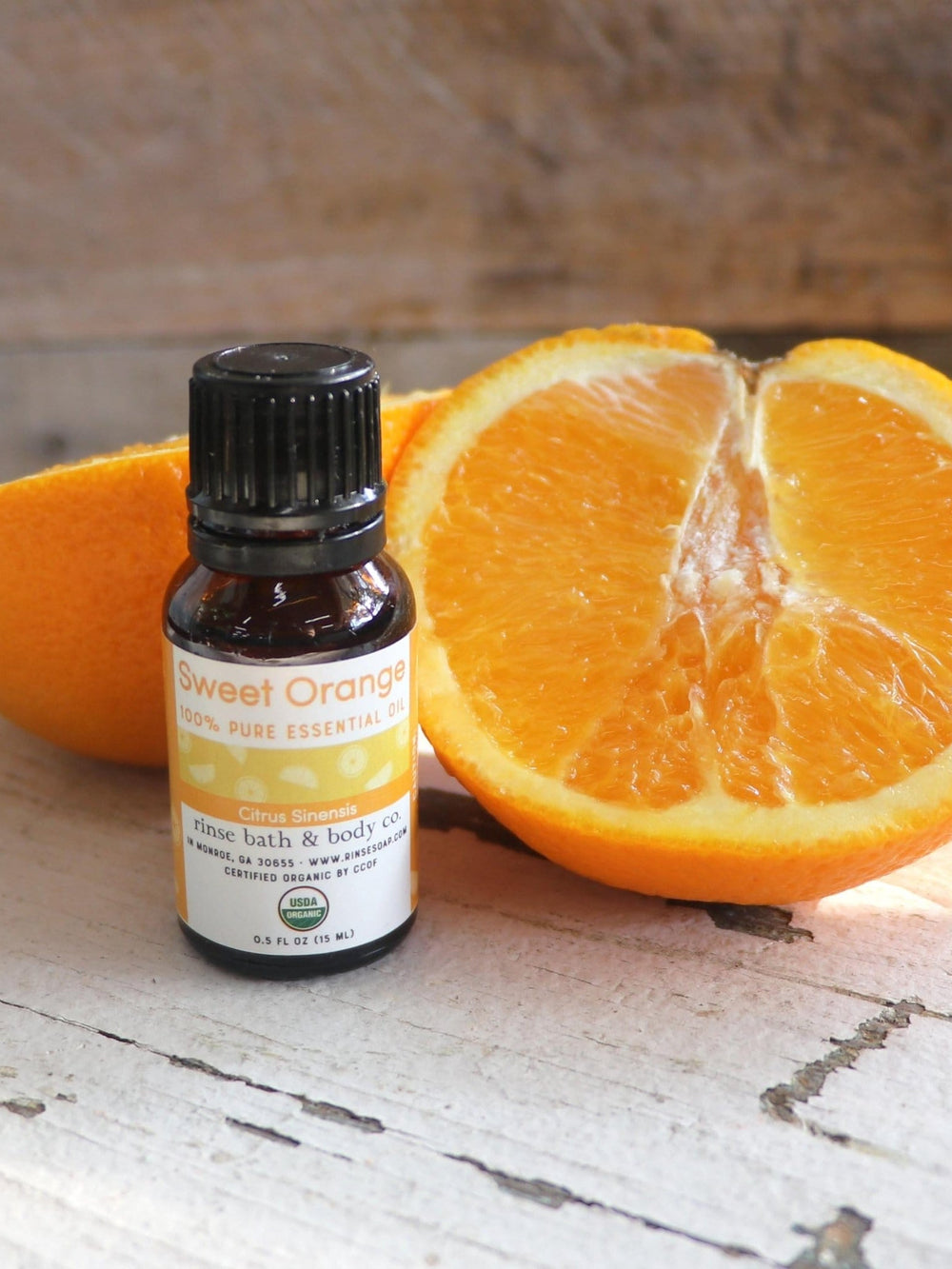 Sweet Orange essential oil standing beside a juicy halved orange on weathered white wood — warm rustic daylight