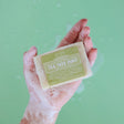 Sudsy hand holding Tea Tree Mint soap — lime label against a mint-green backdrop
