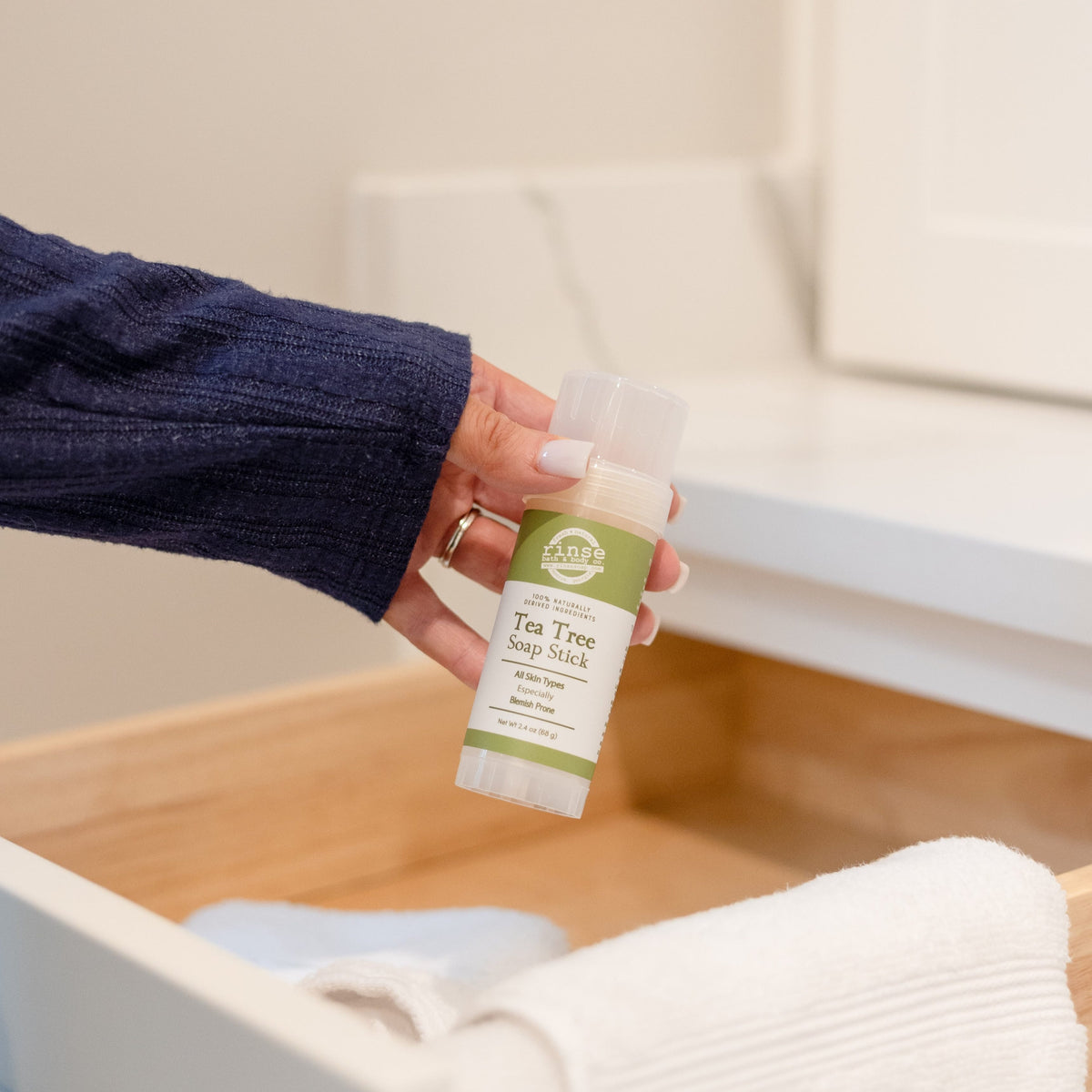 Hand lifting a Tea Tree Soap Stick from an open wooden drawer of white and blue towels — green label in bright light