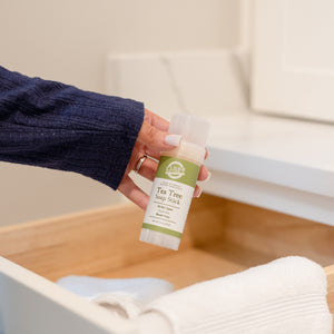 Hand lifting a Tea Tree Soap Stick from an open wooden drawer of white and blue towels — green label in bright light