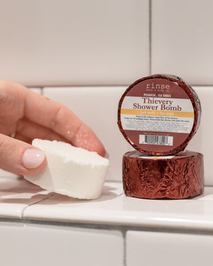 Fingers presenting a textured Thievery shower puck — crinkled burgundy foil on tiled bathroom rim