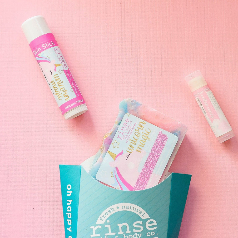 The Unicorn Magic kids gift set on pink linen — swirled unicorn bar soap, Unicorn Magic solid lotion stick and a Cotton Candy lip balm beside the aqua carton. Rinse Bath & Body Co.