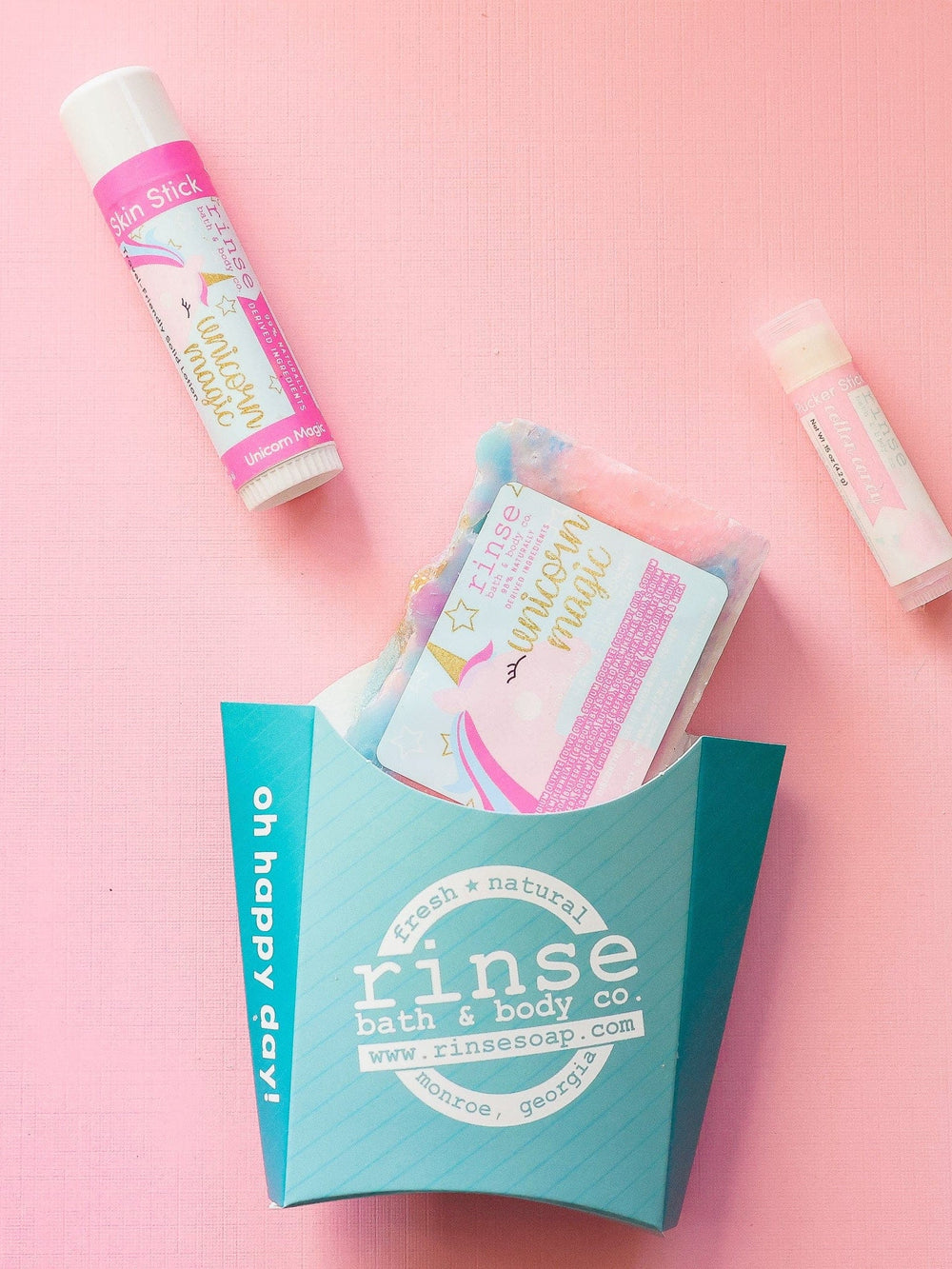 The Unicorn Magic kids gift set on pink linen — swirled unicorn bar soap, Unicorn Magic solid lotion stick and a Cotton Candy lip balm beside the aqua carton. Rinse Bath & Body Co.