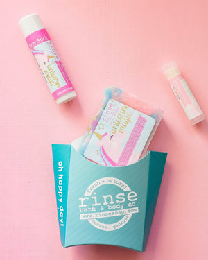 The Unicorn Magic kids gift set on pink linen — swirled unicorn bar soap, Unicorn Magic solid lotion stick and a Cotton Candy lip balm beside the aqua carton. Rinse Bath & Body Co.