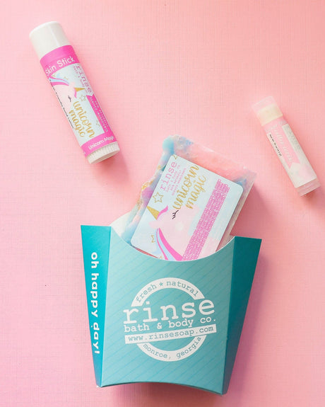 The Unicorn Magic kids gift set on pink linen — swirled unicorn bar soap, Unicorn Magic solid lotion stick and a Cotton Candy lip balm beside the aqua carton. Rinse Bath & Body Co.
