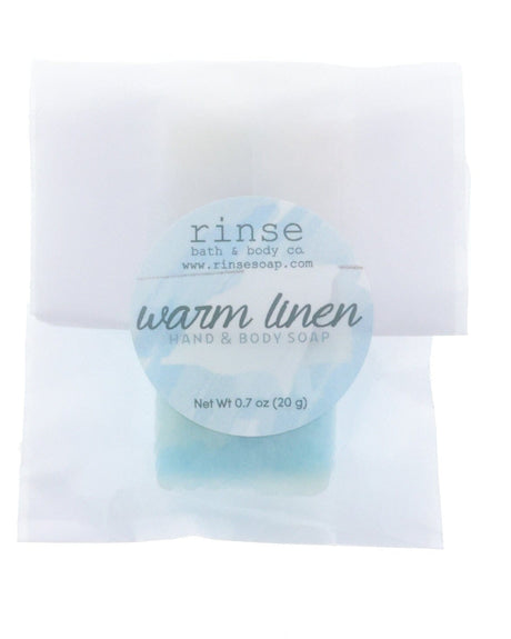 Warm Linen soap slice rests in crinkled translucent paper under a soft blue circular label on bright white.
