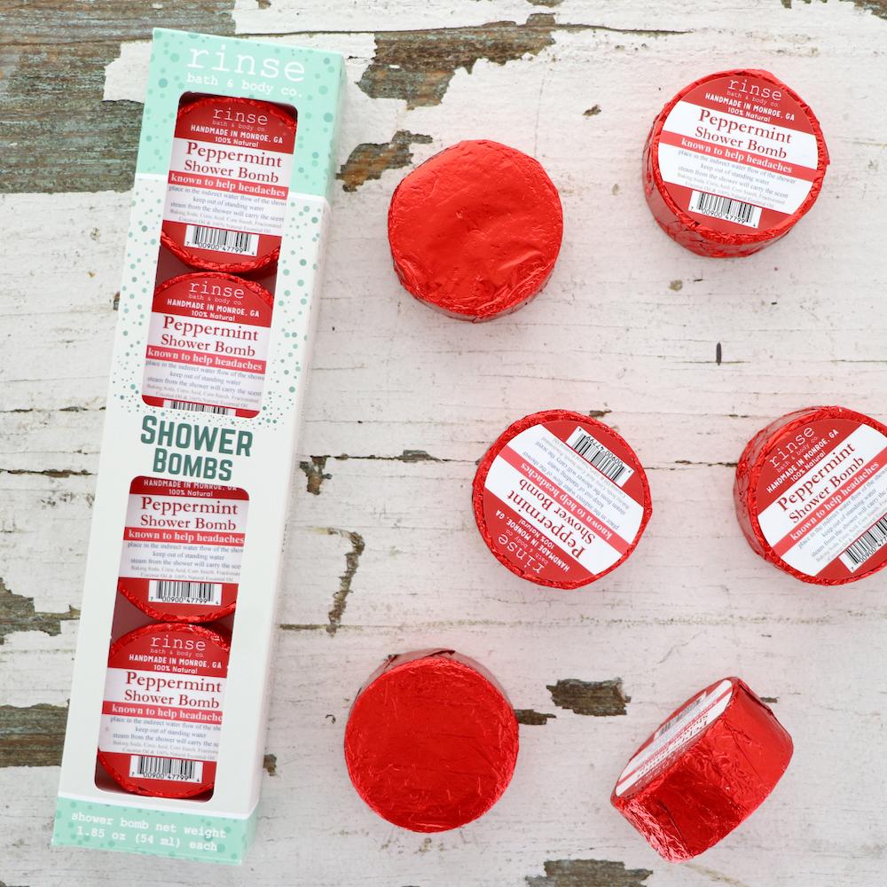 The Peppermint Shower Steamer 4-pack box on chipped white-painted wood with four red foil-wrapped pucks scattered beside it, one unwrapped to show the white steamer — peppermint steam for a blocked head. Rinse Bath & Body Co.