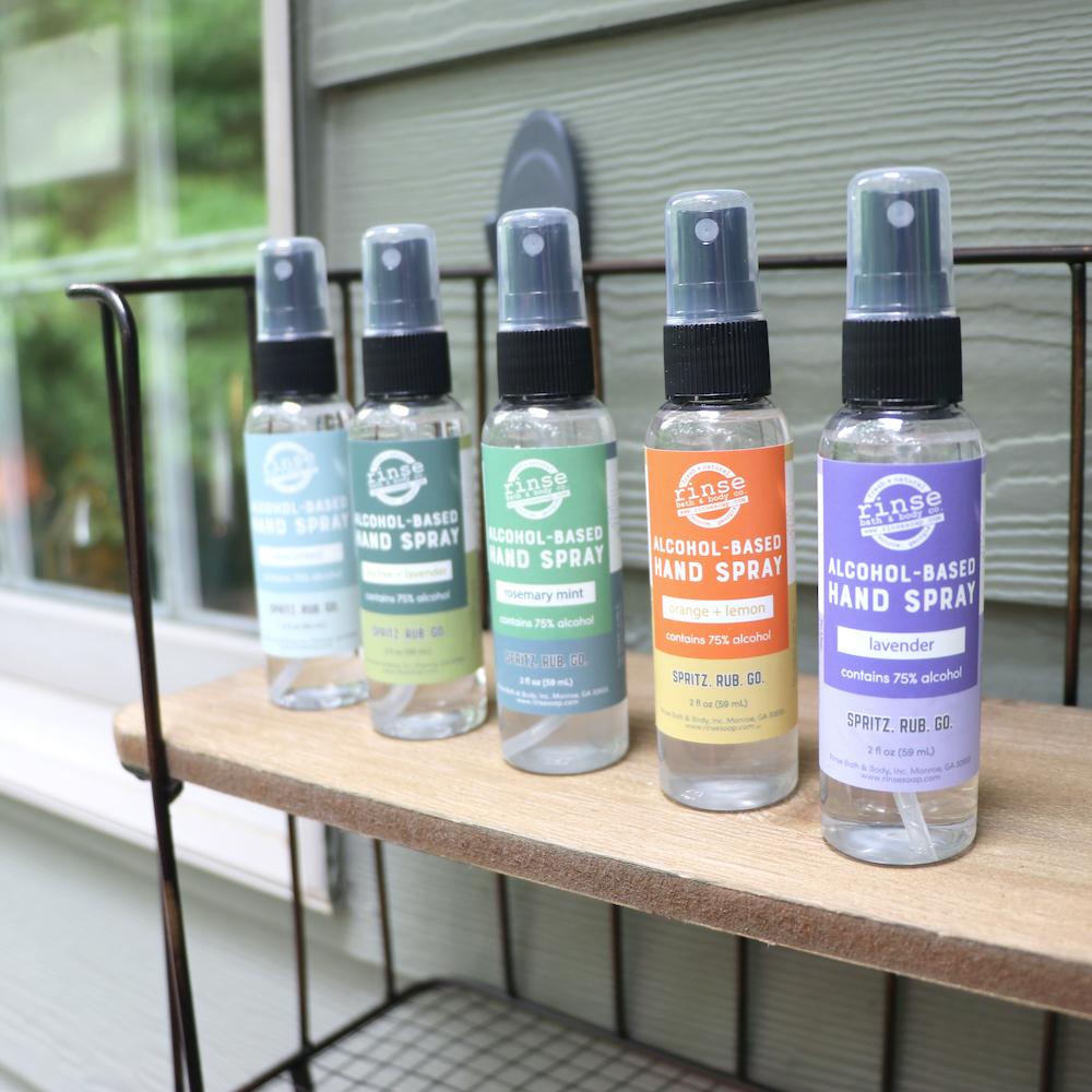 Five colorful hand sprays line a wire wood shelf — sage green siding in soft outdoor light