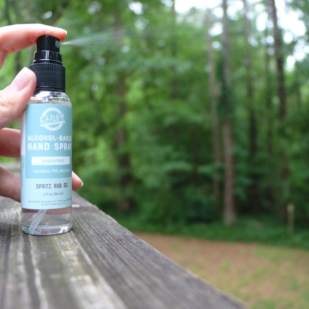 Hand misting Unscented spray on a wooden deck — soft green trees blurred behind in outdoor light